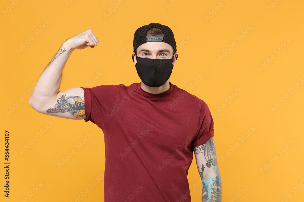 Young tattooed man guy in casual t-shirt cap black face mask posing isolated on yellow background studio portrait. People sincere emotions lifestyle concept. Mock up copy space Showing biceps muscles. Young tattooed man guy in casual t-shirt cap black face mask posing isolated on yellow background studio portrait. People sincere emotions lifestyle concept. Mock up copy space Showing biceps muscles.