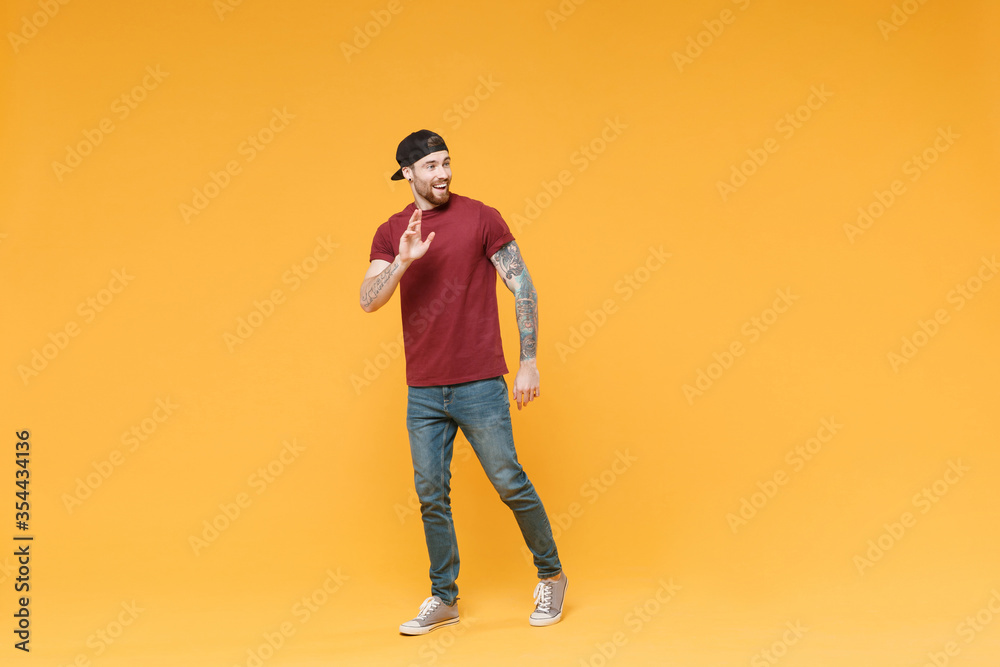 Cheerful young tattooed man guy in casual t-shirt black cap posing isolated on yellow background studio. People lifestyle concept. Mock up copy space. Waving and greeting with hand as notices someone. Cheerful young tattooed man guy in casual t-shirt black cap posing isolated on yellow background studio. People lifestyle concept. Mock up copy space. Waving and greeting with hand as notices someone.