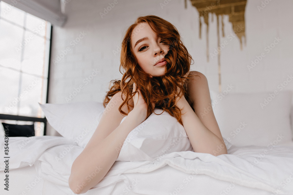 Debonair dark-haired woman lying on white sheet. Indoor photo of cheerful relaxed girl with ginger hair.