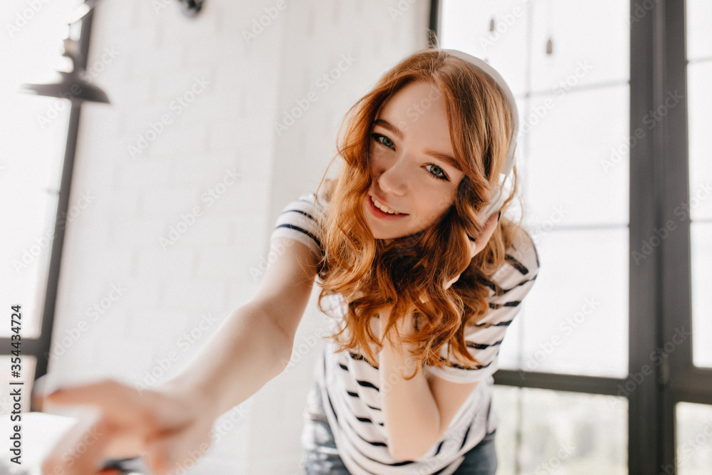 Good-humoured girl in headphones looking to camera. Charming curly female model having fun at home.