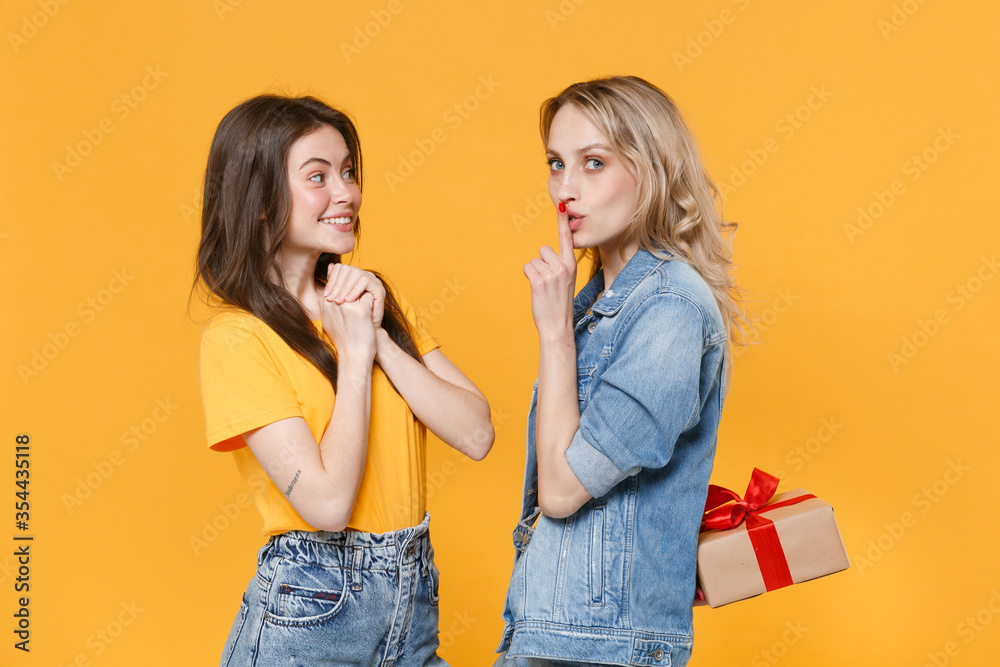 Excited secret women girls friends in denim clothes isolated on yellow background. Birthday, holiday concept. Hold present box with gift ribbon bow, say hush be quiet with finger on lips shhh gesture. Excited secret women girls friends in denim clothes isolated on yellow background. Birthday, holiday concept. Hold present box with gift ribbon bow, say hush be quiet with finger on lips shhh gesture.