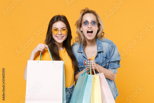 Smiling amazed young women girls friends in denim clothes eyeglasses posing isolated on yellow background. People sincere emotions lifestyle concept. Holding package bag with purchases after shopping.