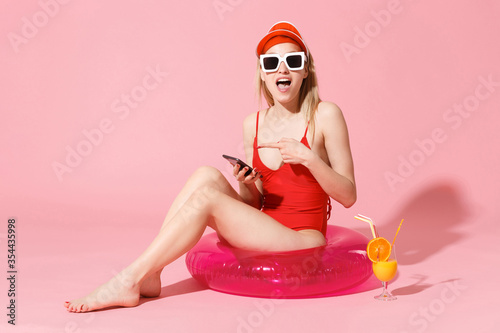 Shocked young blonde woman girl in red one-piece swimsuit cap sunglasses isolated on pink background. People summer vacation rest lifestyle concept. Sit in swim inflatable ring, point on mobile phone.