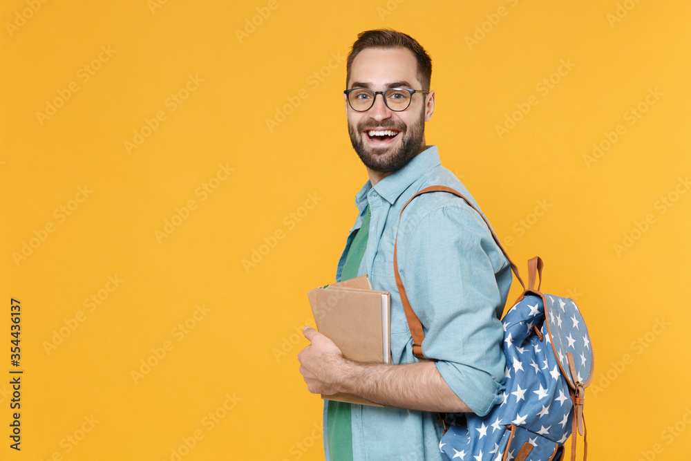 Fototapeta premium Side view of funny man student in casual clothes glasses backpack hold books isolated on yellow background. Education in high school university college concept. Mock up copy space. Looking camera.