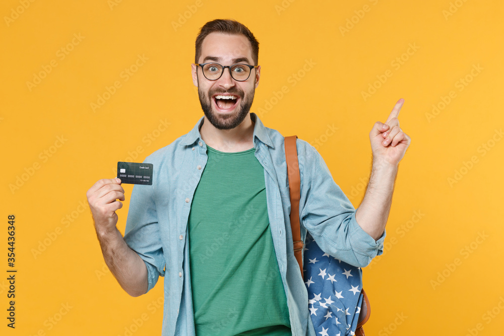 Excited young man student in casual clothes glasses with backpack isolated on yellow background. Education in high school university college concept. Hold credit bank card pointing index finger up. Excited young man student in casual clothes glasses with backpack isolated on yellow background. Education in high school university college concept. Hold credit bank card pointing index finger up.