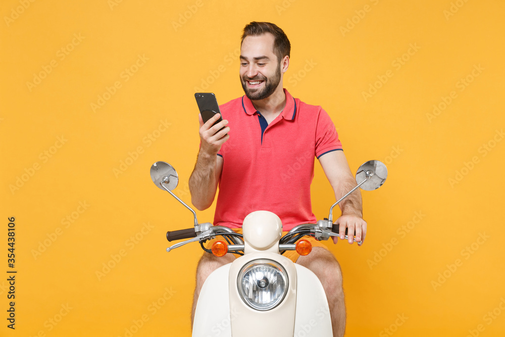 Smiling young bearded guy in casual summer clothes driving moped isolated on yellow background. Driving motorbike transportation concept. Mock up copy space. Using mobile phone, typing sms message.