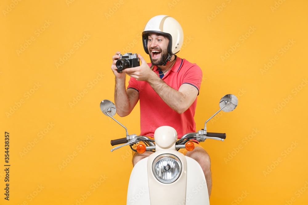 Funny young bearded man guy in casual summer clothes helmet driving moped isolated on yellow background. Driving motorbike transportation concept. Mock up copy space. Hold retro vintage photo camera.