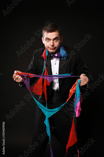 Funny magic man in tuxedo and bow tie with colorful handkerchief isolated on black background