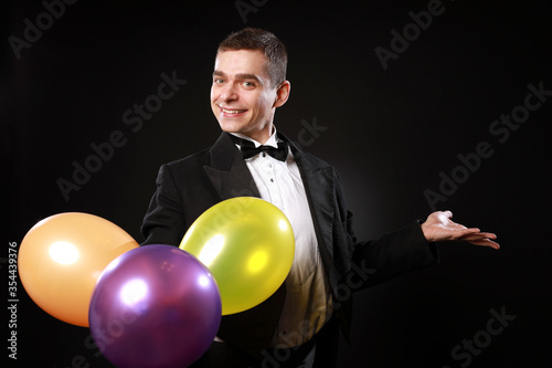 Magic man in tuxedo and bow tie with colorful balloons isolated on black background © Naz