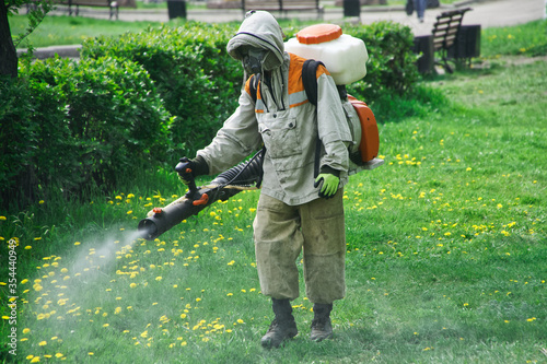 A worker in a protective suit and respirator performs sanitary and chemical treatment in a park or park. Spraying a repellent or antiviral compound on the street photo