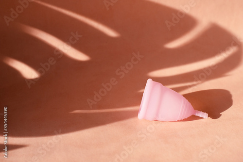 Menstrual cup on a pink background with the shadow of a tropical palm leaf. The concept of critical days during the holidays and summer