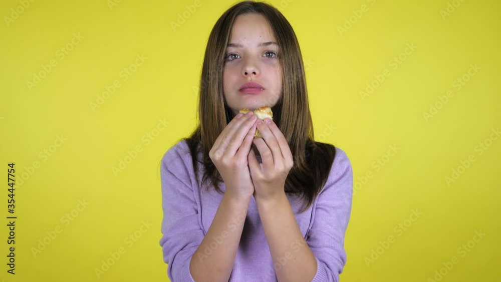 Teen girl in a pink sweater bites and eats burger on yellow background with copy space. Place for text or product. 4K slow motion footage Teen girl in a pink sweater bites and eats burger on yellow background with copy space. Place for text or product. 4K slow motion footage