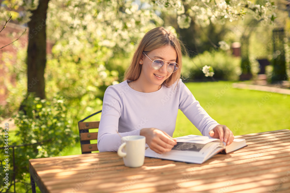 Obraz premium A cute young teenage girl is reading a book while sitting at a wooden table with a Cup of coffee, in the back yard.