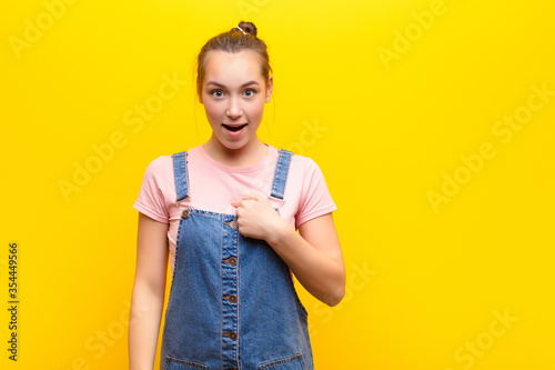 young blonde pretty girl looking shocked and surprised with mouth wide open, pointing to self against yellow wall