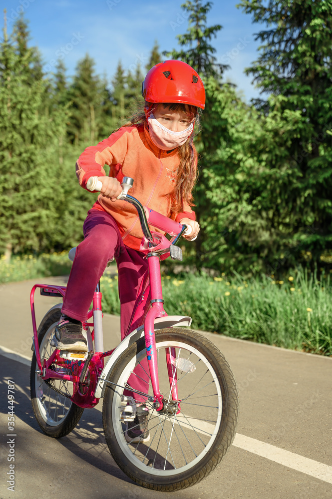 Cute little girl in a protective mask rides a bicycle in a city park. Vertical orientation. Cute little girl in a protective mask rides a bicycle in a city park. Vertical orientation.