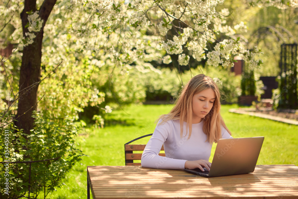 A cute young teen girl is learning online on a laptop while sitting at a wooden table in the back garden of a house. A cute young teen girl is learning online on a laptop while sitting at a wooden table in the back garden of a house.