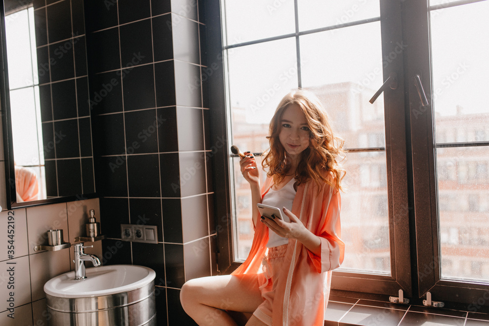 Romantic curly woman with phone doing skincare routine. Indoor photo of slim white girl with makeup brush. - obrazy, fototapety, plakaty 