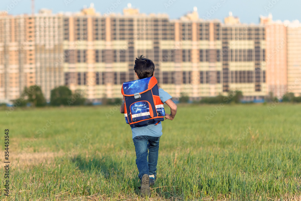 The schoolboy happily runs with a backpack on the field against the background of the city landscape