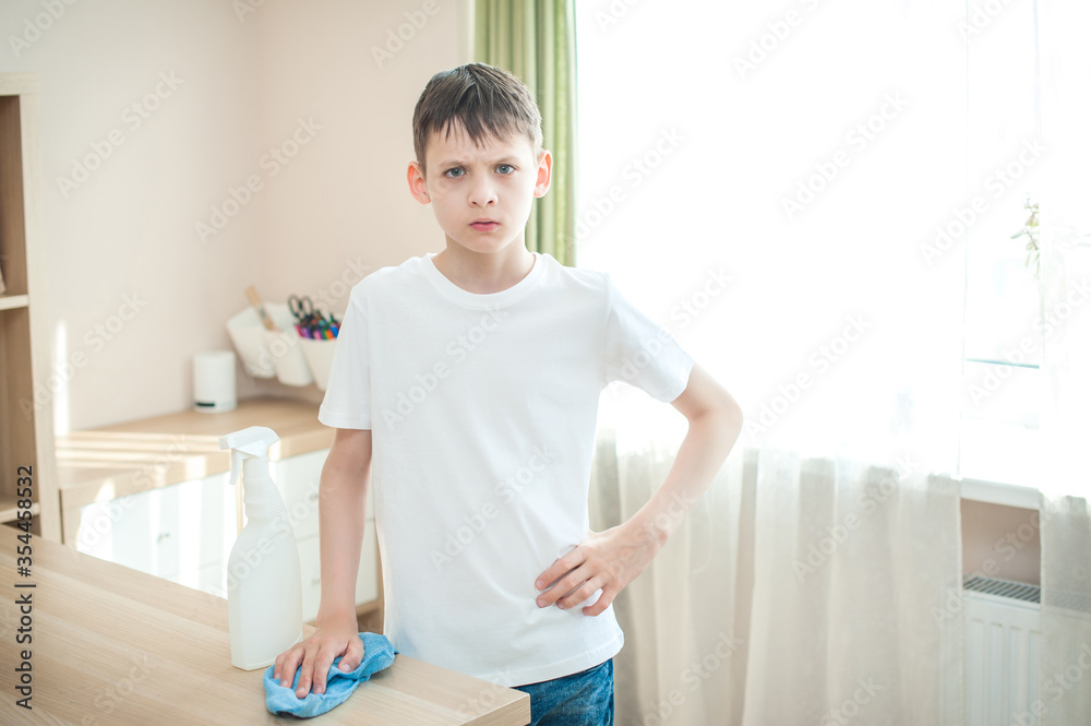 A teenager in jeans and a white T-shirt is standing near the table. He does not want to wipe the table, angry. A rag in hand and a cleaning agent nearby. A teenager in jeans and a white T-shirt is standing near the table. He does not want to wipe the table, angry. A rag in hand and a cleaning agent nearby.