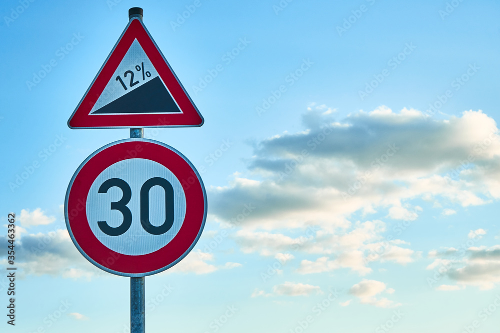 A red and white traffic sign, which warns of a twelve percent gradient and a traffic sign below it with a speed limit of 30 km/h. A blue sky with white clouds is in the background.