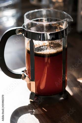 Glass coffee cafetiere (also known as a french press) filled with freshly brewed black coffee grounds on a kitchen counter in the morning sun.