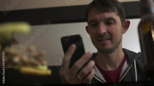 High quality slow motion close-up 4k video of a young blurred man chatting with friend or family on phone watching tv and having snacks with bottle of beer. Happy time. Handsome person with phone. 