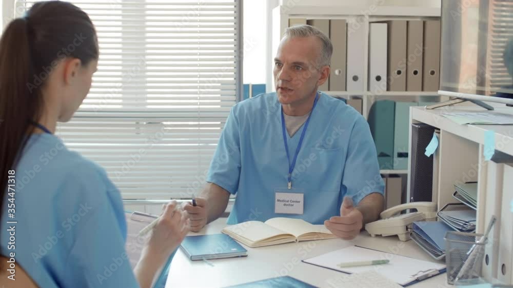 Rear view of middle-aged male doctor consulting female colleague with dark hair sitting in front of him and making notes in chart