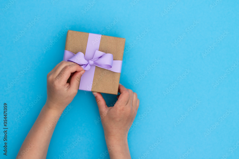 Hands receiving a gift in craft paper with a lilac ribbon on blue background. Top view. Copy space. Hands receiving a gift in craft paper with a lilac ribbon on blue background. Top view. Copy space.