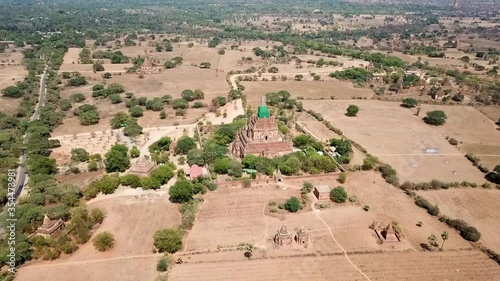 Aerial shot of Buddhist temple amidst plants and field, drone flying forward towards pagodas on sunny day - Bagan, Myanmar photo