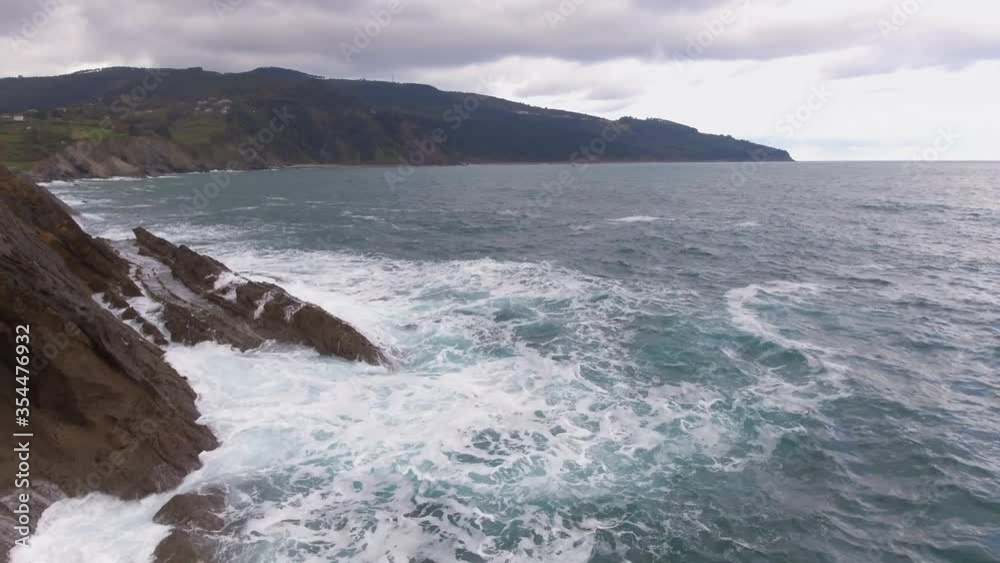 Retreating drone shot of a giant rock structure amidst the sea; waves crashing ashore; mountain shore in BG Retreating drone shot of a giant rock structure amidst the sea; waves crashing ashore; mountain shore in BG