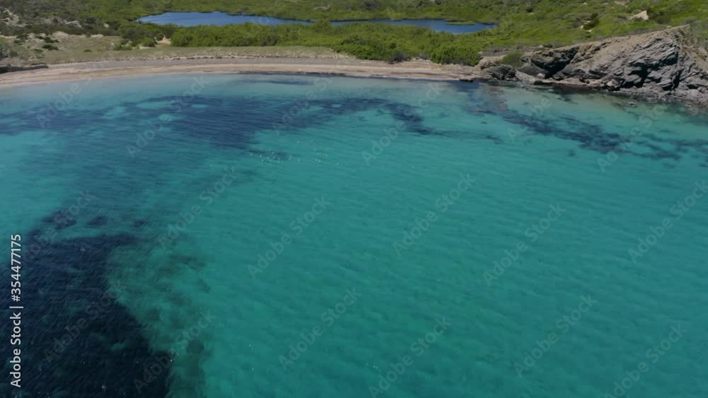 Crystal Clear Blue Water Of Ocean With Wetlands And Green Mountain Hills On The Background On A Sunny Summer Day In Menorca, Balearic Islands, Spain, - aerial drone shot Crystal Clear Blue Water Of Ocean With Wetlands And Green Mountain Hills On The Background On A Sunny Summer Day In Menorca, Balearic Islands, Spain, - aerial drone shot