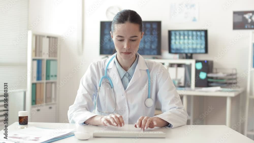 Medium shot portrait of female Caucasian virologist wearing medical overall sitting at computer, typing on wireless keyboard then looking at camera and smiling Medium shot portrait of female Caucasian virologist wearing medical overall sitting at computer, typing on wireless keyboard then looking at camera and smiling
