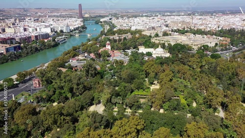 Aerial shot of green trees in park by river on sunny day, drone flying forward towards university against sky - Seville, Spain