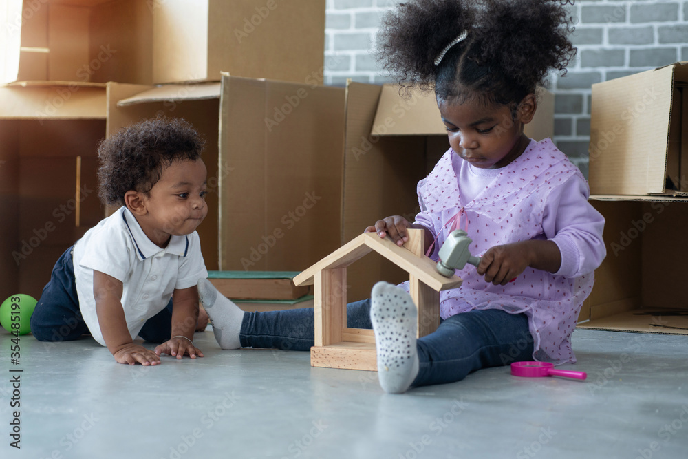 Dark skinned baby boy crawled on the floor and watching his sister playing in wooden house, stacks of cardboard boxes in background Dark skinned baby boy crawled on the floor and watching his sister playing in wooden house, stacks of cardboard boxes in background