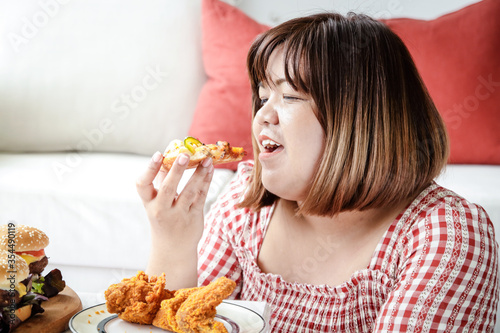 An Asian fat woman is eating pizza and fried chicken on the sofa in her house. The concept of consuming food that causes disease is not good for health