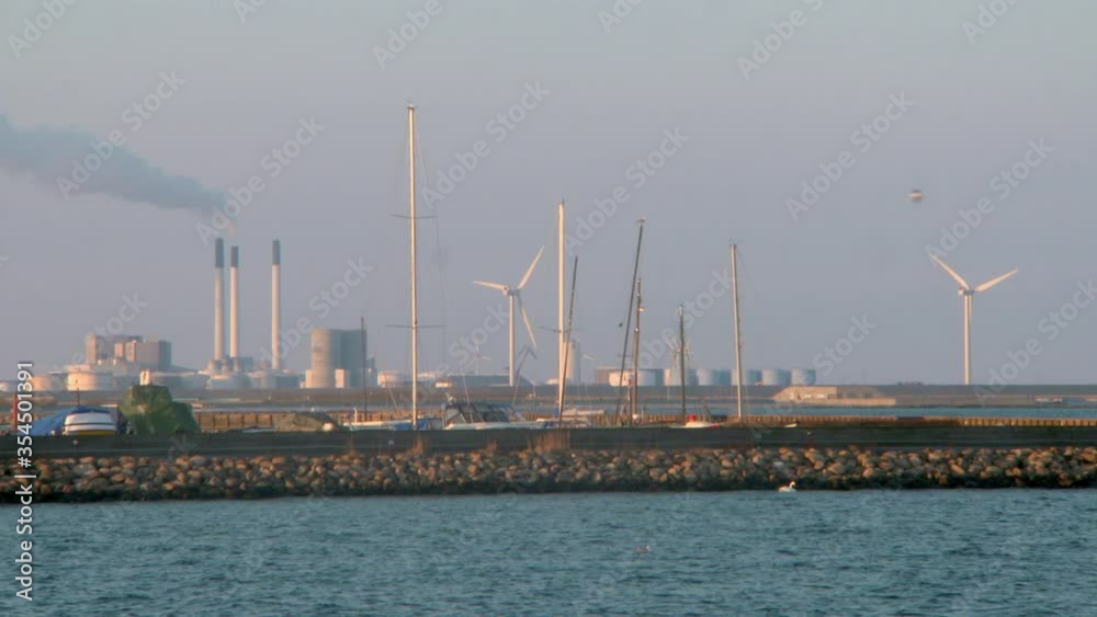 Lockdown shot of wind turbines and factory by sea against sky - Copenhagen, Denmark Lockdown shot of wind turbines and factory by sea against sky - Copenhagen, Denmark