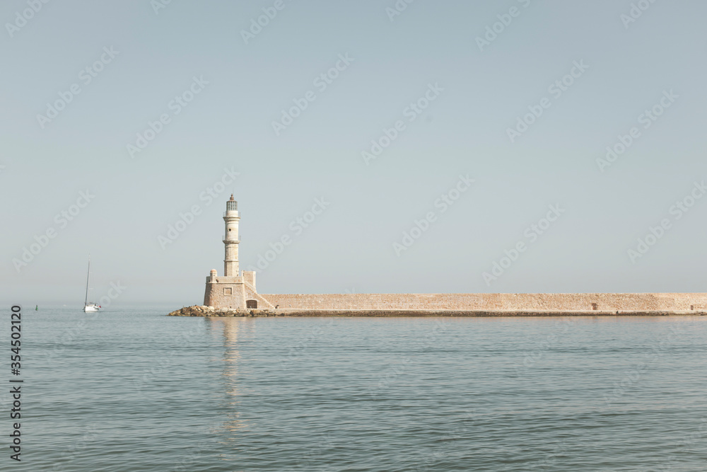 An old lighthouse in the middle of the sea, a yacht is sailing in the distance. Calm and blue sky