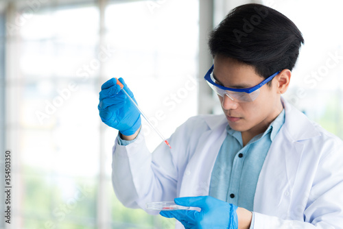 Professional Asian scientist or researcher man in work coat uniform do experiment pipetting red liquid sample to mix in petri dish in science lab