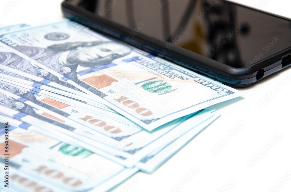 Dollars and smartphone. Banknotes in denominations of 100 dollars lie on a white background on them, next lies a smartphone with a black screen. Top view and angle. Macro smartphone and dollars Dollars and smartphone. Banknotes in denominations of 100 dollars lie on a white background on them, next lies a smartphone with a black screen. Top view and angle. Macro smartphone and dollars