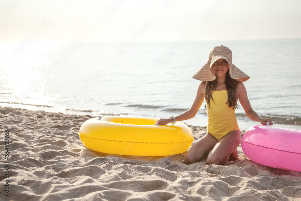 Tan girl wearing straw hat and yellow swimsuit relax on the beach and take sunbath on summer vacation. child sit on the towel on the sand beach and eat the fruit at the background sea and rubber ring Tan girl wearing straw hat and yellow swimsuit relax on the beach and take sunbath on summer vacation. child sit on the towel on the sand beach and eat the fruit at the background sea and rubber ring