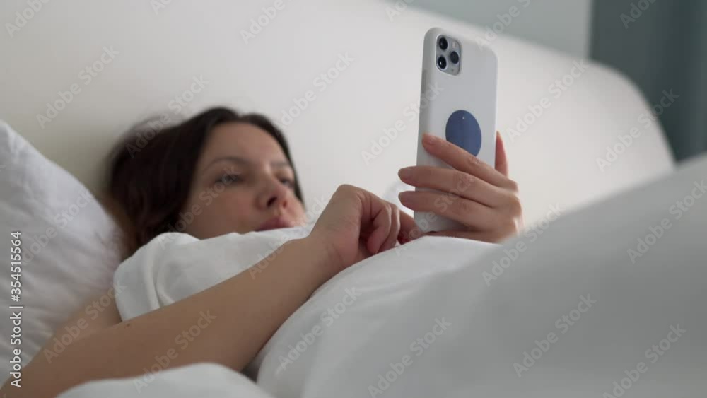 Woman holding phone lying awake in bed in the morning, using social media apps texting on smartphone in bedroom. Woman holding phone lying awake in bed in the morning, using social media apps texting on smartphone in bedroom.