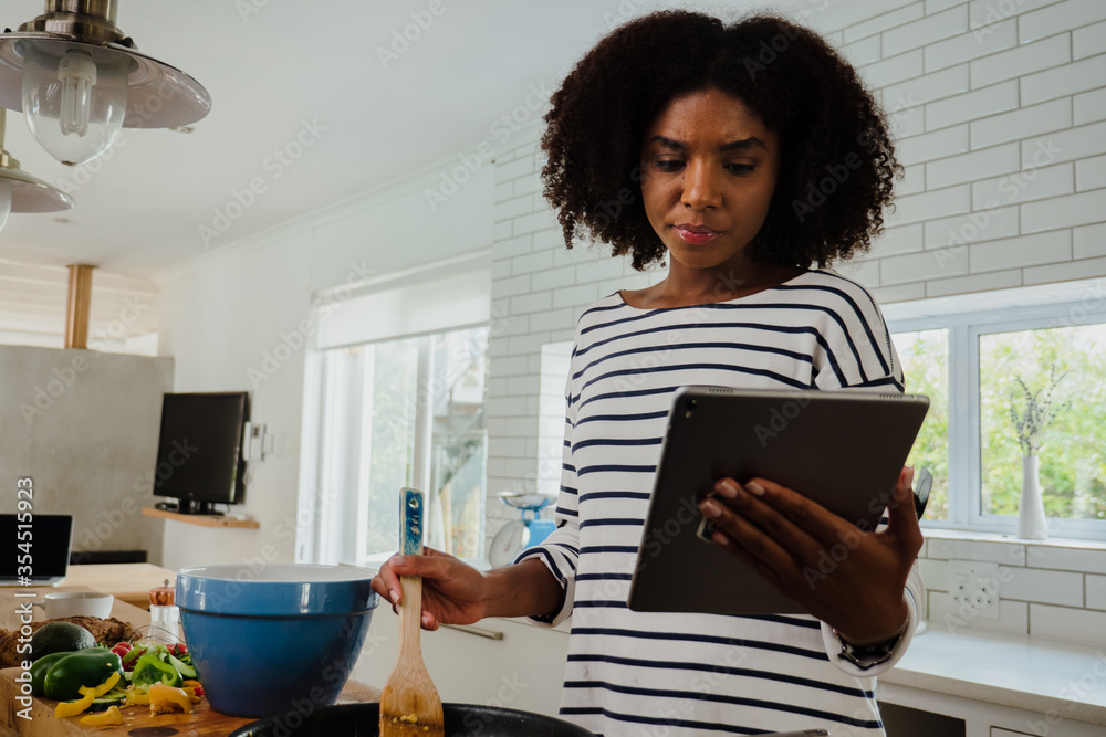 Beautiful mixed race woman looking at recipe on tablet while cooking in her kitchen at home Beautiful mixed race woman looking at recipe on tablet while cooking in her kitchen at home