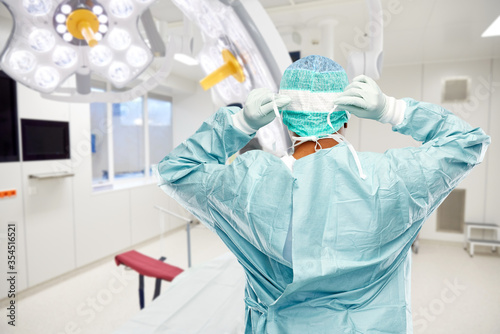 medicine  surgery and people concept - male doctor or surgeon in protective wear putting face mask on over operating room background