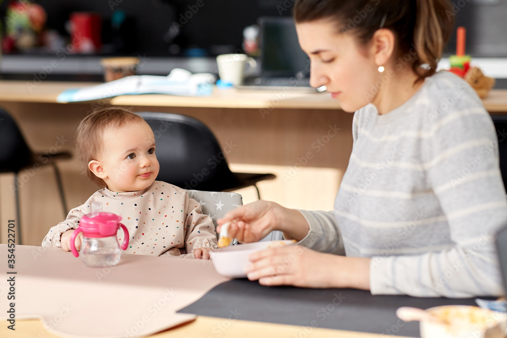family, eating and people concept - happy mother with puree and spoon feeding little baby girl sitting in highchair at home family, eating and people concept - happy mother with puree and spoon feeding little baby girl sitting in highchair at home
