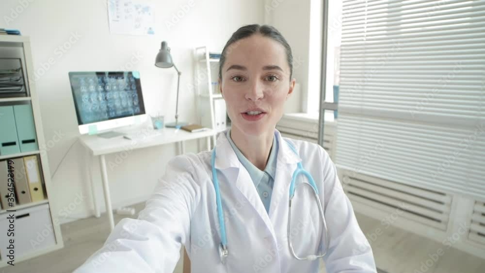 Medium shot of Caucasian female medical worker wearing medical overall and stethoscope holding camera and prescribing red pill to online patient Medium shot of Caucasian female medical worker wearing medical overall and stethoscope holding camera and prescribing red pill to online patient