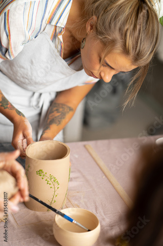 Smiling beautiful girl in blue apron holding rolling pin and happily working with clay at pottery class studio. male potter master rolling up the clay on table with ceramic products.