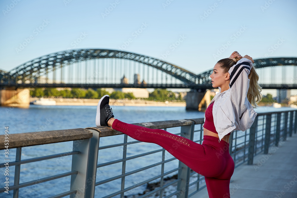 Fit in the city, young woman doing gymnastics and stretching exercises on the river early in the morning after sunrise