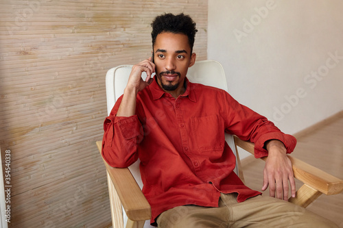 Bewildered young short haired bearded guy with dark skin looking attentively in front of himself while making call with his mobile phone, sitting in cosy chair over beige interior