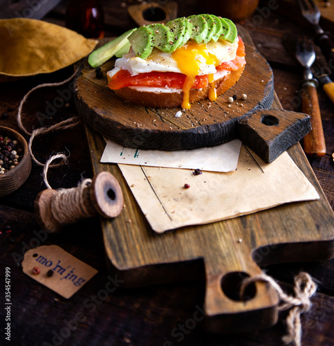 tasty open sandwich or toast with poached egg, salmon and sliced avocado on wooden boards stands on rustic table