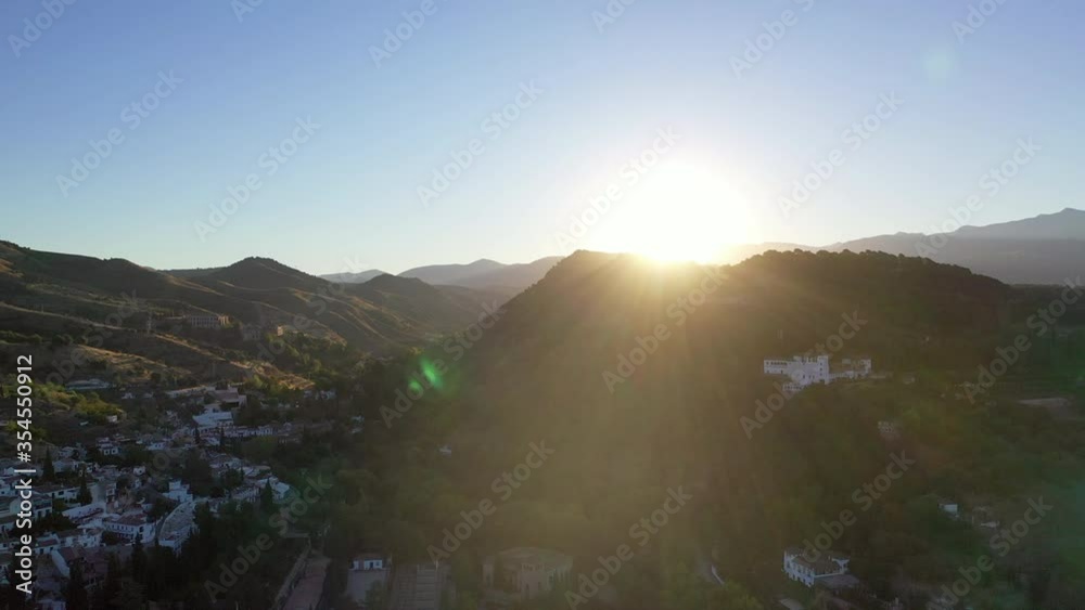 Aerial shot of buildings by mountains against sky during sunset, drone flying backward from sun over mountain range - Granada, Spain Aerial shot of buildings by mountains against sky during sunset, drone flying backward from sun over mountain range - Granada, Spain